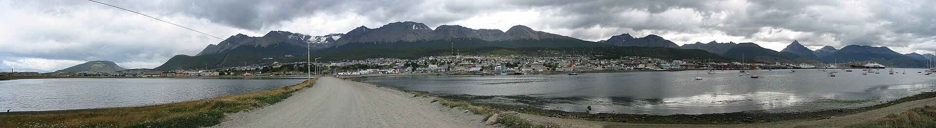 A small city across a gray waterway under lowering gray clouds. A road leads to the city across a causeway. Mountains with snow and a low treeline form the backdrop. A few boats are in the water.